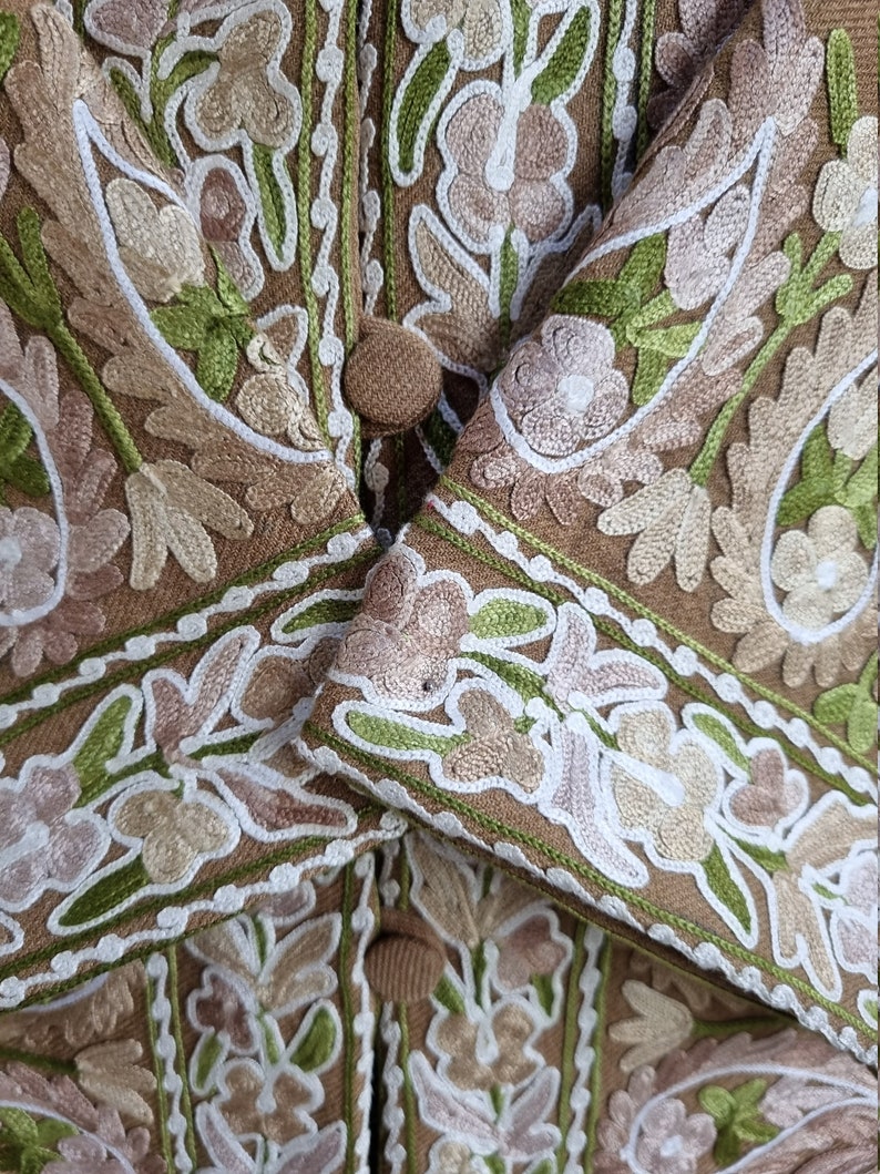K&ouml;nnte beinhalten: Nahaufnahme einer braunen Stoffjacke mit aufwendigen Stickereien in Gr&uuml;n-, Wei&szlig;- und Rosat&ouml;nen. Die Jacke hat ein Blumenmuster und einen Knopfverschluss.