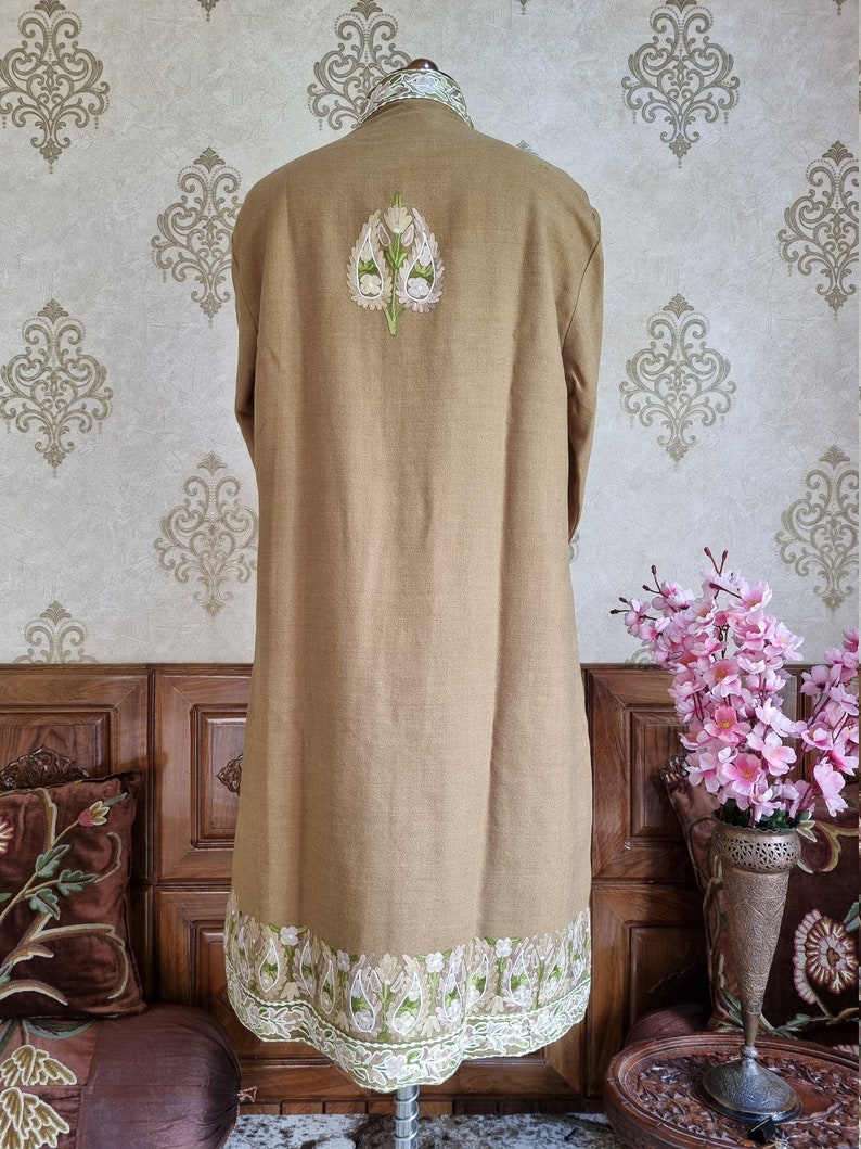 K&ouml;nnte beinhalten: Ein langer, brauner Leinenmantel mit wei&szlig;en und gr&uuml;nen Stickereien. Die Stickerei zeigt ein florales Design mit einem zentralen Motiv einer stilisierten Blume.