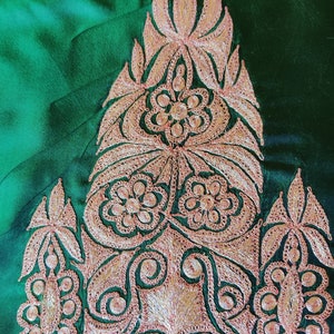 May include: A close-up of a green fabric with intricate gold embroidery. The design features a central motif of a stylized flower with leaves and smaller floral details.