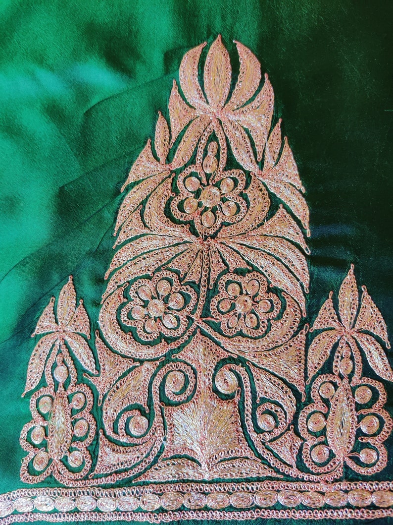 May include: A close-up of a green fabric with intricate gold embroidery. The design features a central motif of a stylized flower with leaves and smaller floral details.