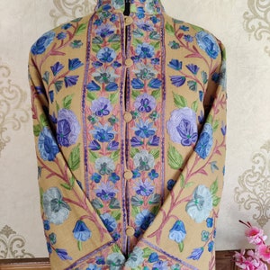 May include: A long, tan-colored coat with a mandarin collar and floral embroidery in shades of blue, purple, and green. The coat has a button closure and long sleeves.