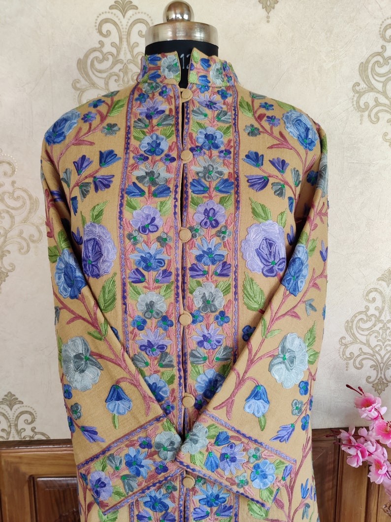 May include: A long, tan-colored coat with a mandarin collar and floral embroidery in shades of blue, purple, and green. The coat has a button closure and long sleeves.