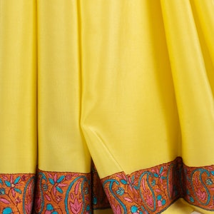 May include: Close-up of a bright yellow textile with a decorative border. The border features a repeating paisley design in shades of orange, pink, and teal. The fabric is draped, showing folds and texture.