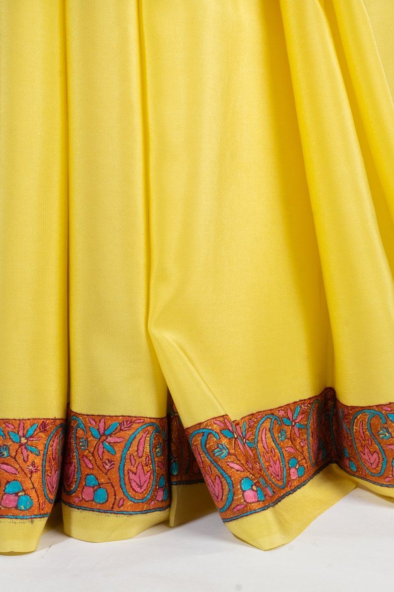 May include: Close-up of a bright yellow textile with a decorative border. The border features a repeating paisley design in shades of orange, pink, and teal. The fabric is draped, showing folds and texture.