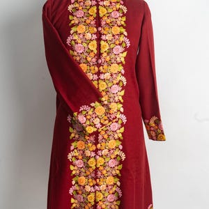Può includere: Una tunica a maniche lunghe color bordeaux intenso con collo alto e un pannello centrale verticale di intricati ricami floreali nei toni del giallo, arancione e rosa. Anche i polsini e l'orlo sono decorati con motivi floreali coordinati.