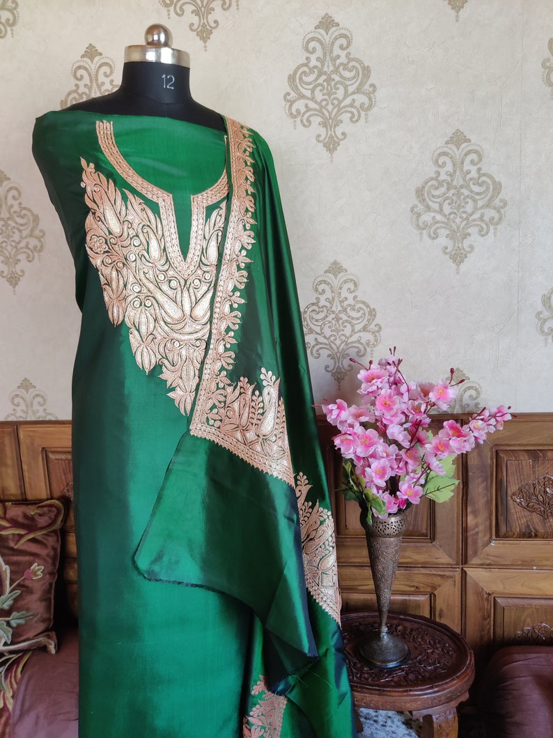 May include: A green silk dress with gold embroidery featuring a floral and bird design. The dress is displayed on a mannequin and has a scoop neckline.