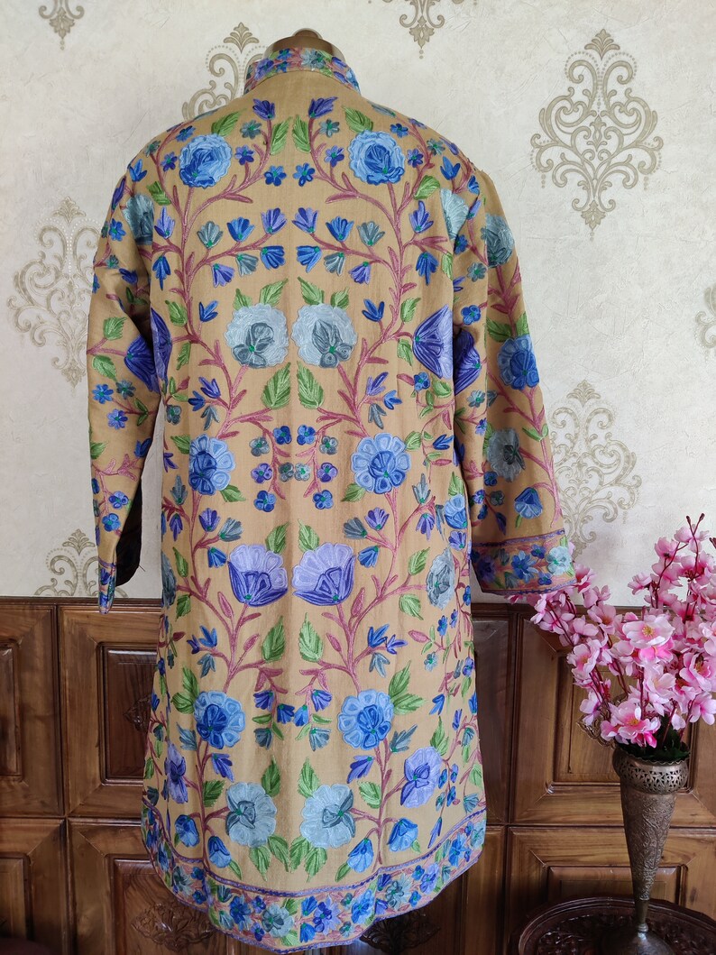 May include: A long, beige embroidered jacket with a floral pattern in shades of blue, green, and pink. The jacket has a mandarin collar and long sleeves.
