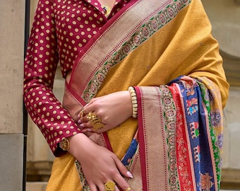 May include: A mustard yellow saree with a wide maroon border featuring intricate green and gold patterns, paired with a long-sleeved maroon blouse with gold polka dots. The ensemble includes a gold choker, dangling earrings, and a headpiece.