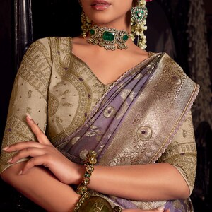 May include: A woman wearing a traditional Indian sari with intricate gold embroidery and a lavender and gold border. She is adorned with emerald and gold jewelry, including a necklace, earrings, and a bracelet. Her hair is styled with flowers.