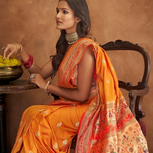 May include: A woman wearing a bright orange and red saree with a floral pattern. The saree has a gold border and is draped over a red blouse. The woman is sitting on a wooden chair and holding a bowl of yellow flowers.