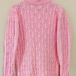 Könnte beinhalten: Ein rosa gestrickter Pullover mit hohem Kragen und einem zarten Zopfmuster.