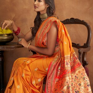 May include: A woman wearing a bright orange and red saree with intricate gold embroidery. The saree has a paisley pattern and a border with floral and bird motifs.