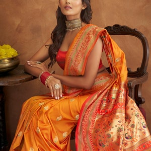 May include: A woman wearing a bright orange and red saree with a paisley pattern. The saree has a gold border with intricate floral and bird designs. The woman is sitting on a chair with a wooden frame.