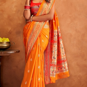May include: A woman wearing a bright orange saree with intricate gold and red embroidery. The saree has a paisley pattern and a border with floral and bird motifs. The woman is wearing a red blouse and gold jewelry.