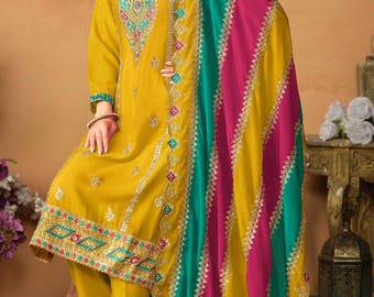 May include: A mustard yellow long-sleeved tunic with intricate embroidery around the neckline and hem, paired with matching trousers. A large, vibrant shawl features alternating vertical stripes of mustard yellow, magenta, and teal with silver trim.