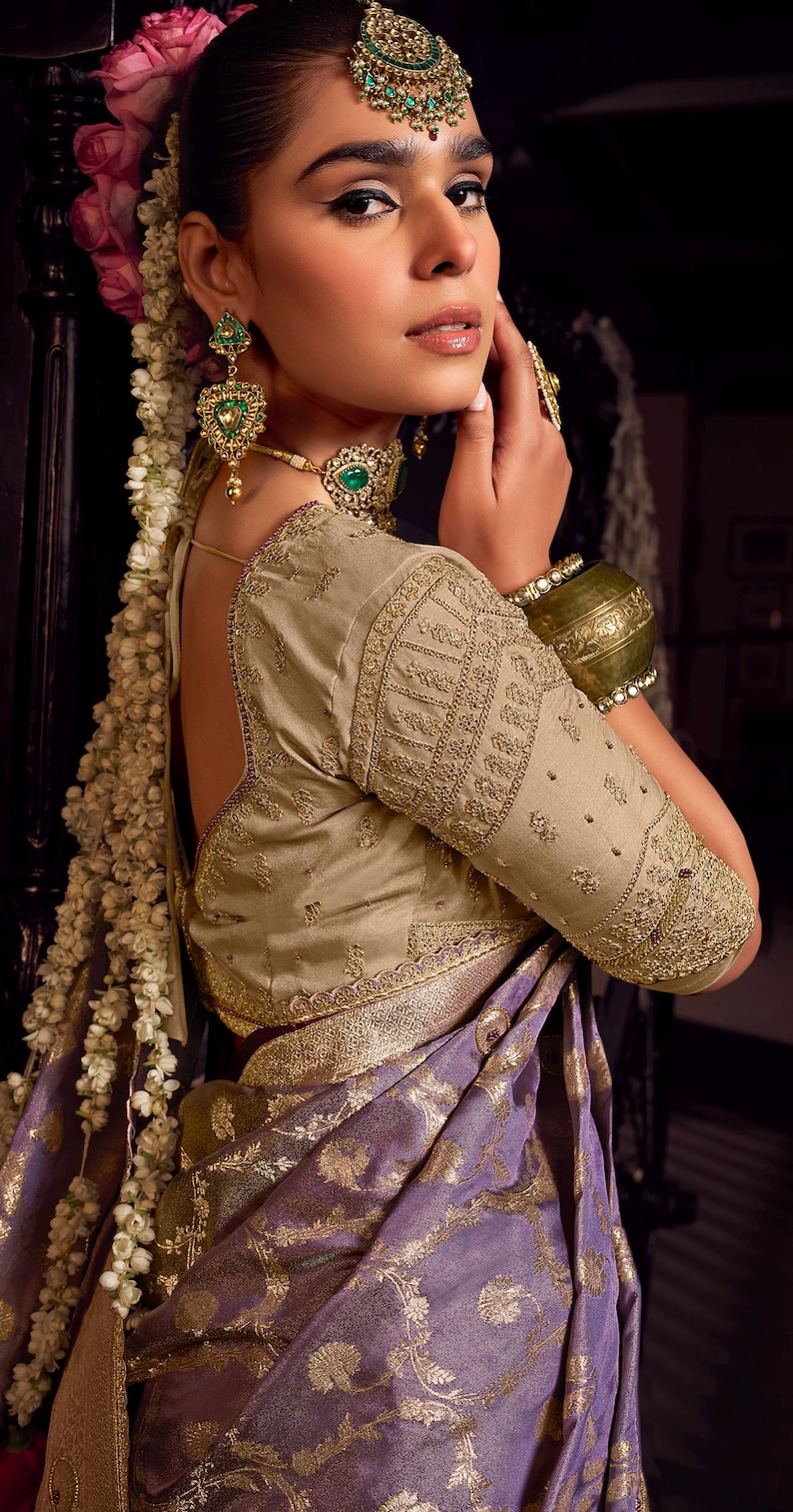 May include: A woman wearing a traditional Indian outfit. The ensemble includes a gold-embroidered blouse, a purple and gold sari, and ornate green and gold jewelry. The woman has flowers in her hair.