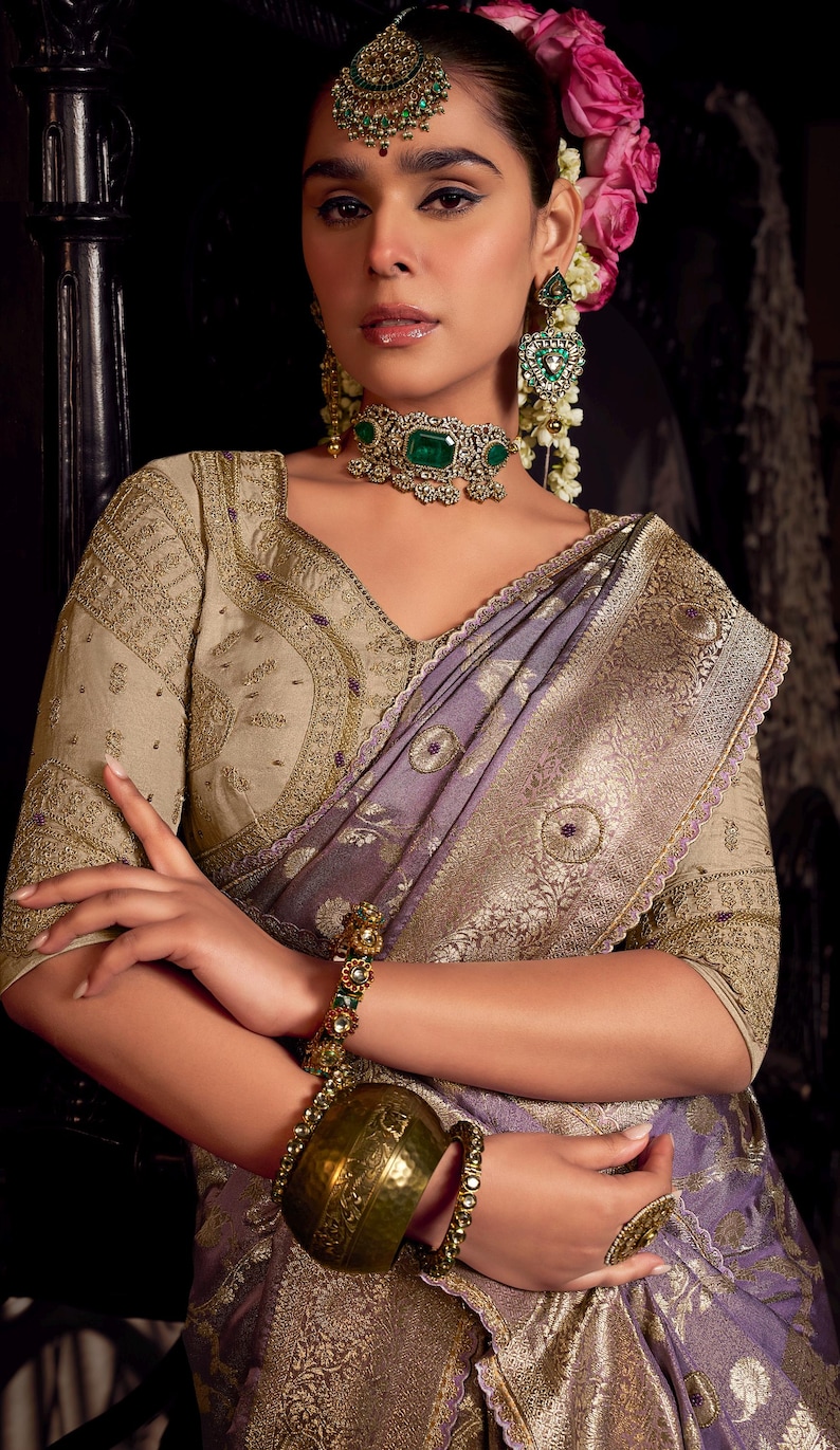 May include: A woman wearing a traditional Indian sari with intricate gold embroidery and a lavender and gold border. She is adorned with emerald and gold jewelry, including a necklace, earrings, and a bracelet. Her hair is styled with flowers.