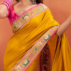 Może przedstawiać: Musztardowe sari ze wzorem z różową obwódką i pasującą bluzką. Sari ma szczegółową obwódkę ze wzorami zwierzęcymi i geometrycznymi w kolorach zielonym, różowym i żółtym. Bluzka ma krótkie rękawy i okrągły dekolt.