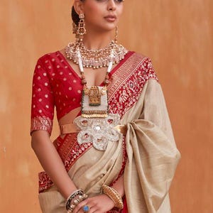 May include: A woman wearing a beige saree with a red and gold border. She is wearing a red blouse with white polka dots. She is also wearing a lot of jewellery, including a large necklace, earrings, bangles, and a headpiece.