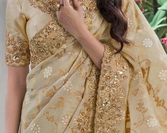 May include: A cream-colored saree with gold embroidery and sequins. The saree has a floral pattern and a gold border. The saree is worn by a woman with long dark hair.
