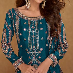 Puede incluir: Ropa formal bordada de color turquesa con intrincados adornos dorados, rosas y plateados. El atuendo de manga larga presenta patrones detallados en el corpiño y las mangas. La modelo lleva pendientes y anillos dorados grandes.