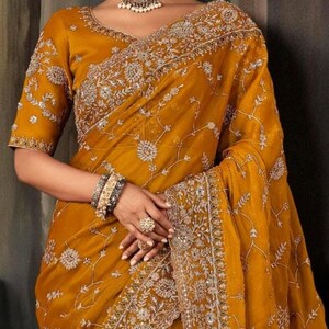 May include: Mustard yellow sari with intricate silver embroidery. The sari features a detailed floral pattern and a decorative border. The outfit includes a matching blouse with short sleeves and a beaded choker necklace and earrings.
