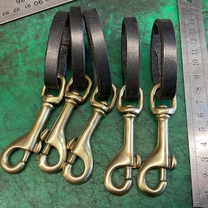 May include: Five brass swivel snap hooks with black leather loops. A metal ruler with centimetre markings is visible, along with a ruler with inch markings. The hooks are on a green surface.
