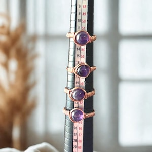 May include: Four amethyst rings with copper wire settings are displayed on a black ring sizer. The rings feature a deep purple gemstone. The copper wire is wrapped around the stone and forms the ring band. The ring sizer has size markings.
