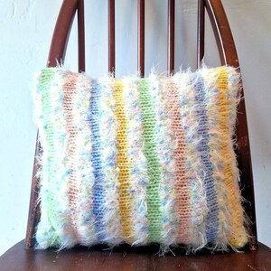 May include: A rectangular, handmade pillow with a striped pattern in pastel colors: yellow, green, pink, and blue. The pillow has a fluffy, textured appearance and is placed on a wooden chair.