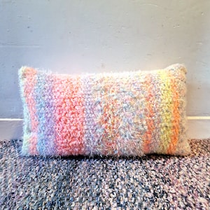 May include: A long, rectangular pillow with a fluffy, fuzzy texture. The pillow is made of a multi-colored yarn with stripes of pink, yellow, purple, and white.