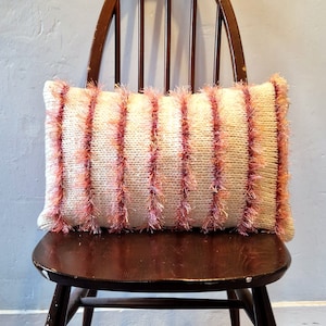 May include: A white and pink striped knitted throw pillow with a fluffy texture. The pillow is sitting on a wooden chair.