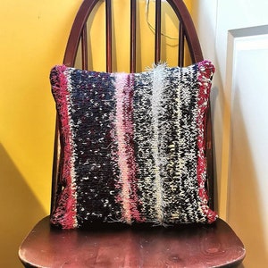 May include: A decorative pillow with a knitted design. The pillow features a mix of black, white, pink, and red yarns. The pillow is rectangular and has a fringe trim along the edges. It is placed on a wooden chair.