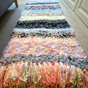 May include: A colorful, handmade rug with a striped pattern. The rug features a variety of colors, including pink, orange, black, white, and blue. The rug has a shaggy texture and a fringe of tassels at the end.