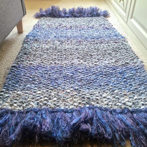 May include: A rectangular rug with a textured weave in shades of gray, blue, and purple. The rug has a fringe of long, shaggy fibers in deep blue and purple at both ends. The rug is placed on a beige carpet.