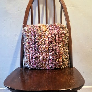 Könnte beinhalten: Ein quadratisches, mehrfarbiges Kissen mit einem strukturierten, geschlungenen Design liegt auf einem dunklen Holzstuhl. Das Kissen hat Farbtöne von Rosa, Weiß, Gelb und Braun. Der Stuhl hat eine geschwungene Rückenlehne und eine dunkle Oberfläche.