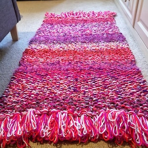 May include: A rectangular, hand-knitted rug in shades of pink, red, and purple. The rug features a fringed edge with white and red yarn. The knitted texture is visible, and the rug appears to be made of thick yarn.