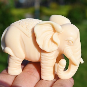 Könnte beinhalten: Eine kleine, elfenbeinfarbene Elefantenfigur mit aufwendigen Details auf dem Rücken. Der Elefant steht auf seinen vier Beinen, sein Rüssel ist eingerollt.