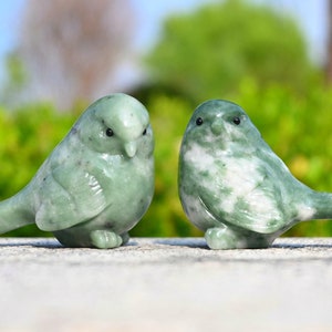 Könnte beinhalten: Zwei grüne Vogelfiguren aus Jadestein. Die Vögel sind realistisch geschnitzt und haben detaillierte Merkmale. Die Vögel schauen sich an und sitzen auf einer grauen Oberfläche.