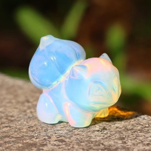 Puede incluir: Una pequeña figurita de un personaje de dibujos animados de ópalo azul iridiscente. La figurita está sentada sobre una superficie gris texturizada.