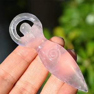 Puede incluir: Un cristal de cuarzo rosa tallado en forma de mujer con un diseño en espiral en la parte delantera. El cristal es translúcido y tiene un acabado liso y pulido.