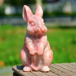 May include: A pink carved rabbit figurine made of polished stone. The rabbit is sitting upright with its paws crossed in front of it.