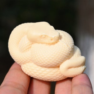 Może przedstawiać: Biała rzeźbiona figurka węża, zwinięta z głową spoczywającą na ciele. Wąż ma szczegółową, fakturowaną powierzchnię.