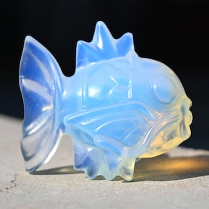 May include: A translucent blue glass sculpture of a fish with a spiky fin and a large eye. The sculpture is made of opalescent glass and has a smooth, polished finish.