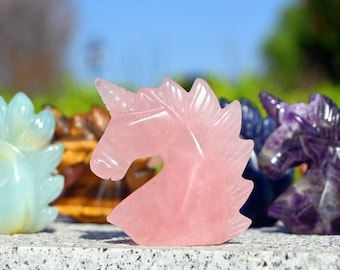 May include: A collection of seven unicorn head carvings, each carved from a different type of crystal. The unicorns are arranged in a row, with the pink rose quartz unicorn in the center.