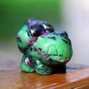 May include: A green and pink carved gemstone figurine of a Pokemon character. The figurine is detailed with black markings and has a shiny, polished finish.
