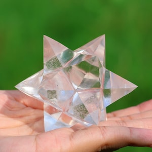 May include: A clear, faceted crystal Merkaba star held in a hand against a blurred green background. The star has multiple triangular faces, catching the light and creating a sparkling effect. The crystal appears to be quartz or a similar transparent material.