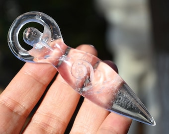 Könnte beinhalten: Eine klare Glasfigur in Form einer stilisierten weiblichen Figur. Die Figur hat einen abgerundeten Kopf mit einem haloartigen Ring darüber, ein Spiralmuster am Torso und eine spitze Basis. Die Figur wird in einer Hand gehalten.