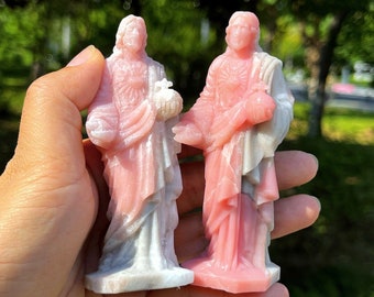 May include: Two small, carved statues of a religious figure, held in a hand. The statues are primarily pink and white, with detailed robes and facial features. The background is blurred, with green foliage.