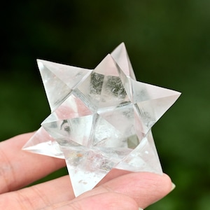May include: A clear, multi-faceted crystal Merkaba star held in a hand. The star has sharp, pointed edges and a geometric design. The background is a blurred green, suggesting an outdoor setting. The crystal is approximately 5 cm across.