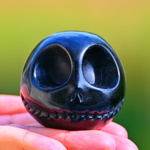May include: A black, spherical sculpture of a character's head with large, oval eye sockets and a stitched mouth. The glossy, polished surface reflects light. The sculpture is held in a hand against a blurred green and yellow background.